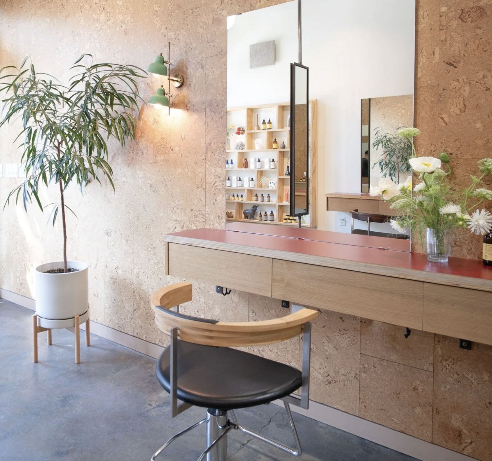 Interior of a modern space with a large potted plant, wooden and metal chair, a cluttered wall-mounted mirror, a wooden surface with a vase of flowers, and a display shelf of bottles and small objects in the background. Head spa certification.