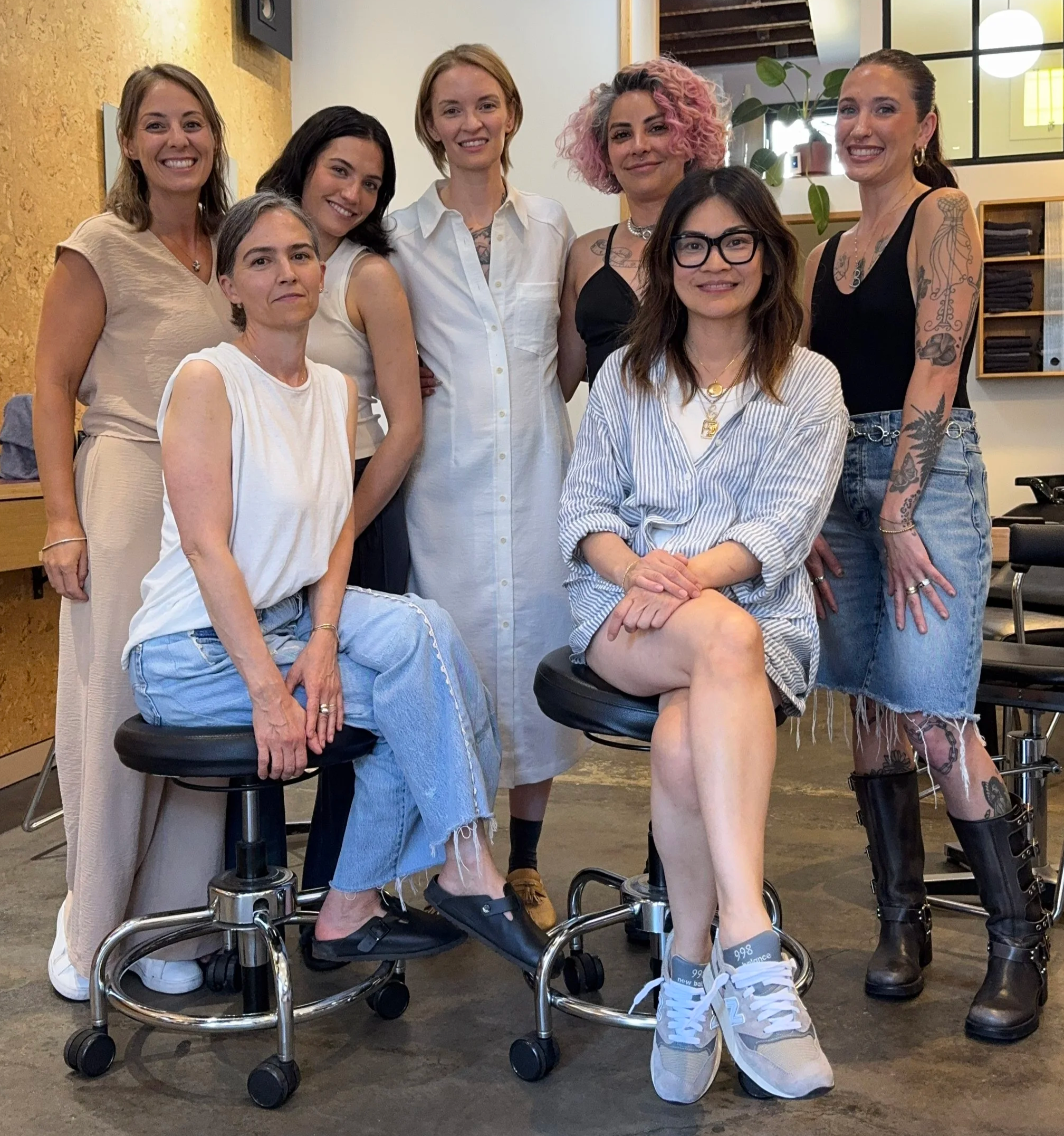 Seven women posing together indoors, some seated on chrome stools, some standing behind, all smiling. The women have diverse styles, including casual, tattooed, and with different hairstyles.  Team training for scalp health.