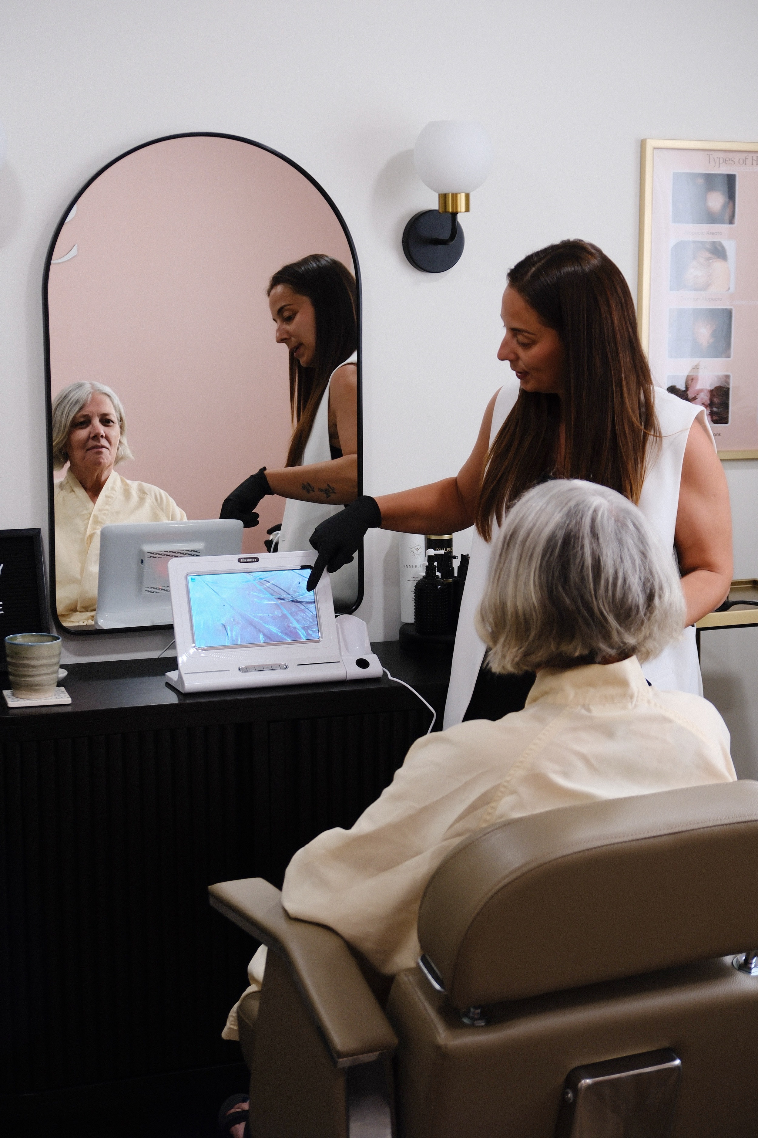 A woman getting her hair done at a salon station, with an older woman seated nearby. The salon has a mirror, wall sconce, and a poster of hair types on the wall. Hair loss and head spa training