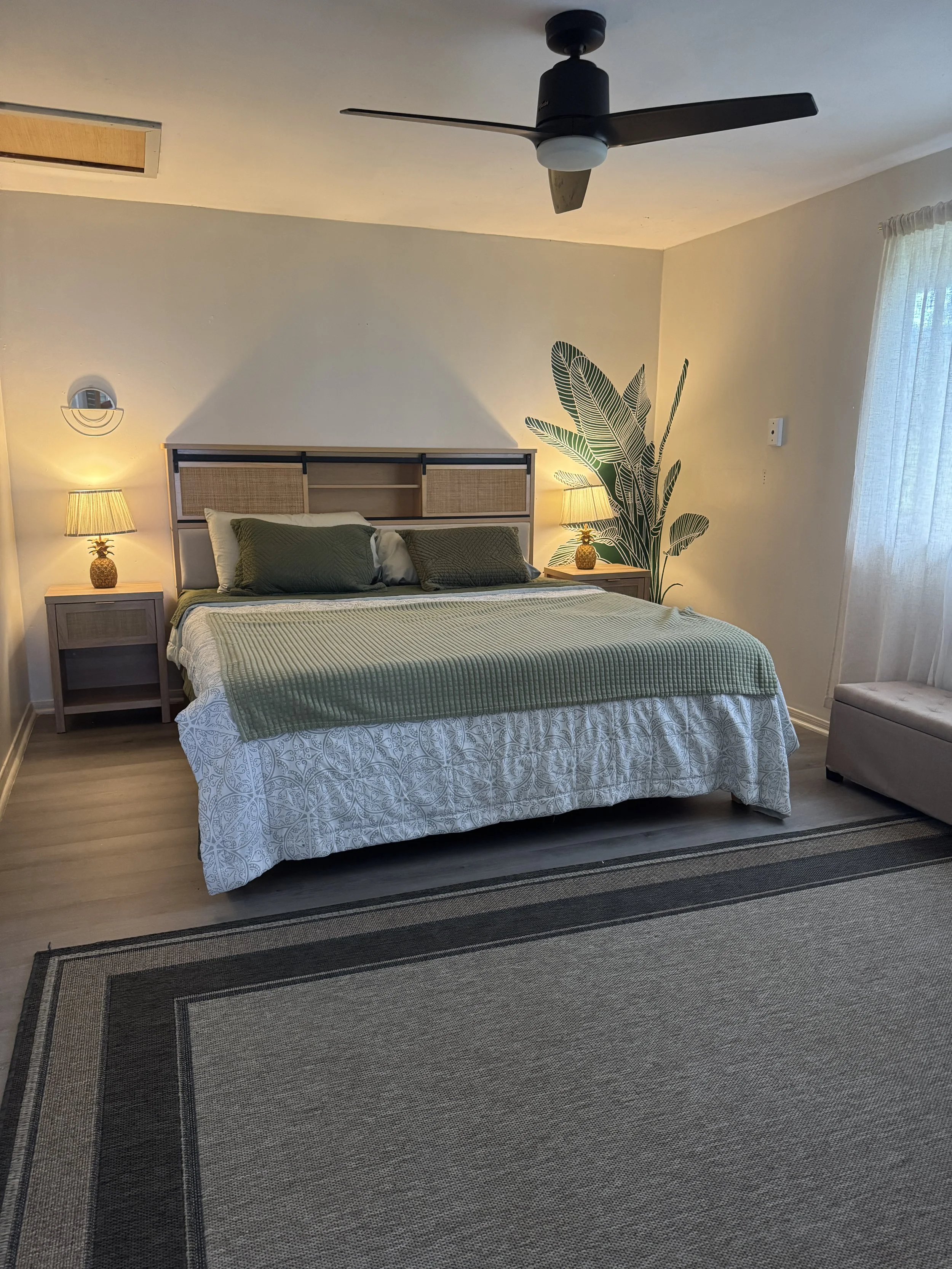 A bedroom with a bed, two nightstands with lamps, a wall mural of large green leaves, a ceiling fan, and a window with curtains.