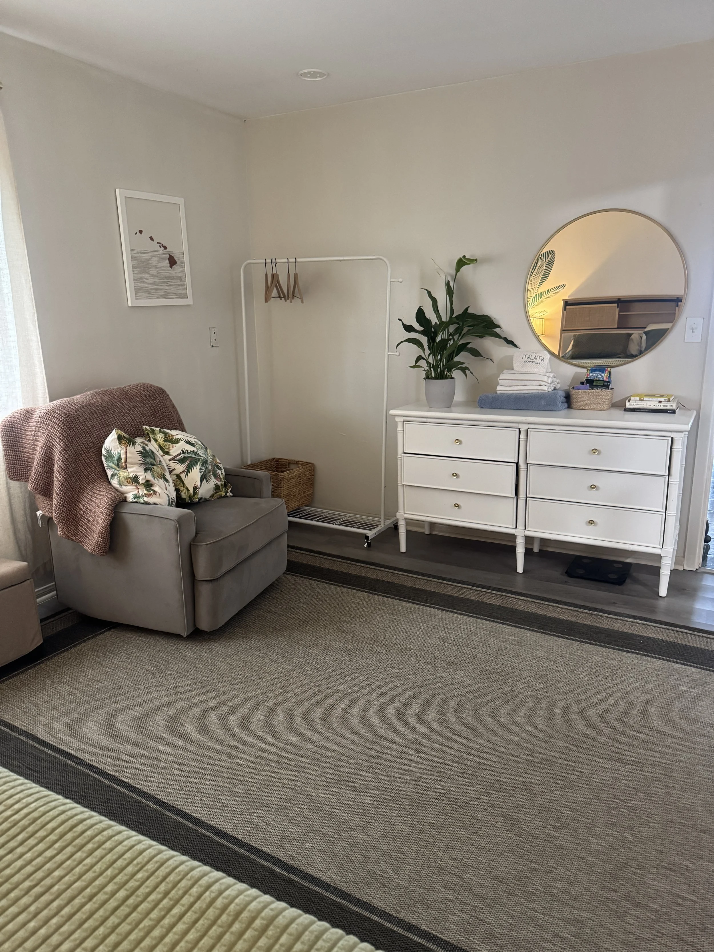 Living room with gray armchair, textured throw blanket, floral pillow, white dresser with stacked towels, potted plant, round mirror, and decorative wall art