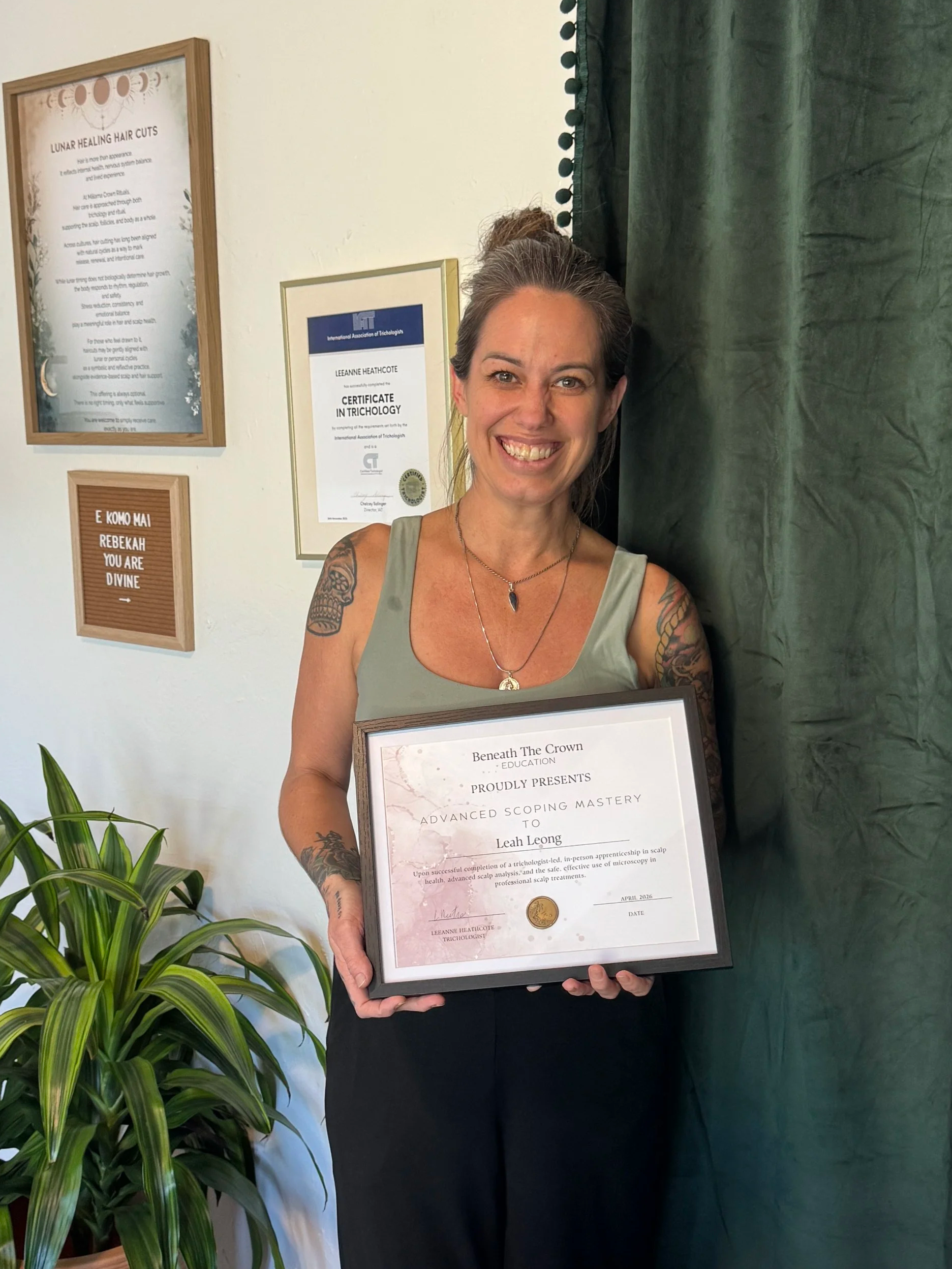Woman holding a framed certificate smiling, with green curtain and framed informational posters on the wall, a potted plant to the left. Scalp care certification.