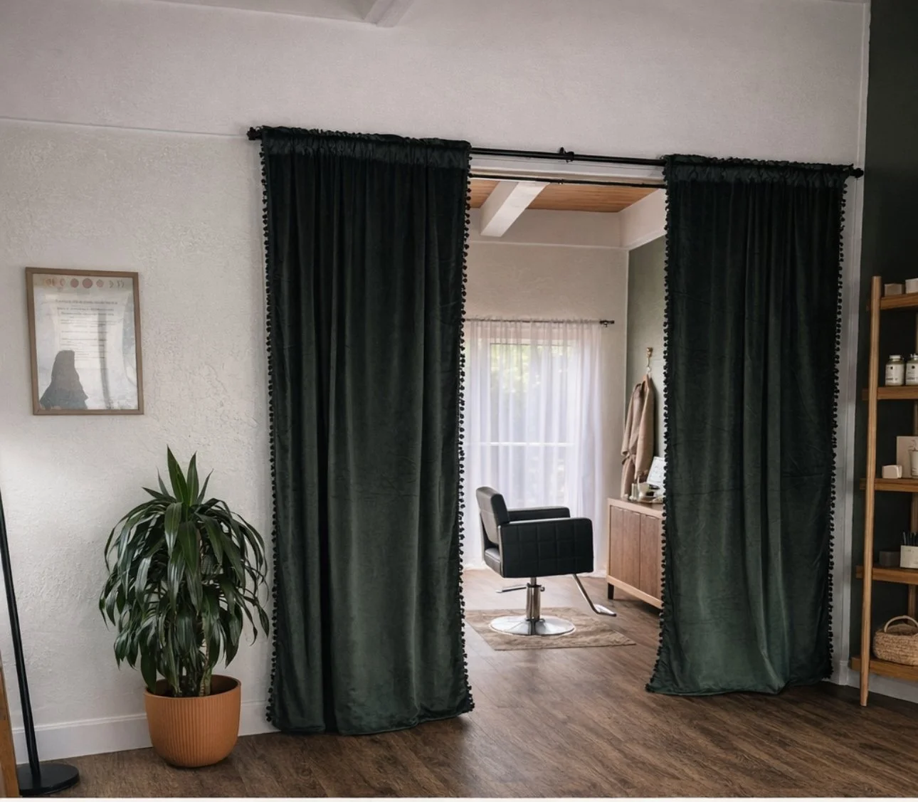 A view through dark green velvet curtains into a room with a black salon chair, a wooden cabinet, and a window with sheer curtains.