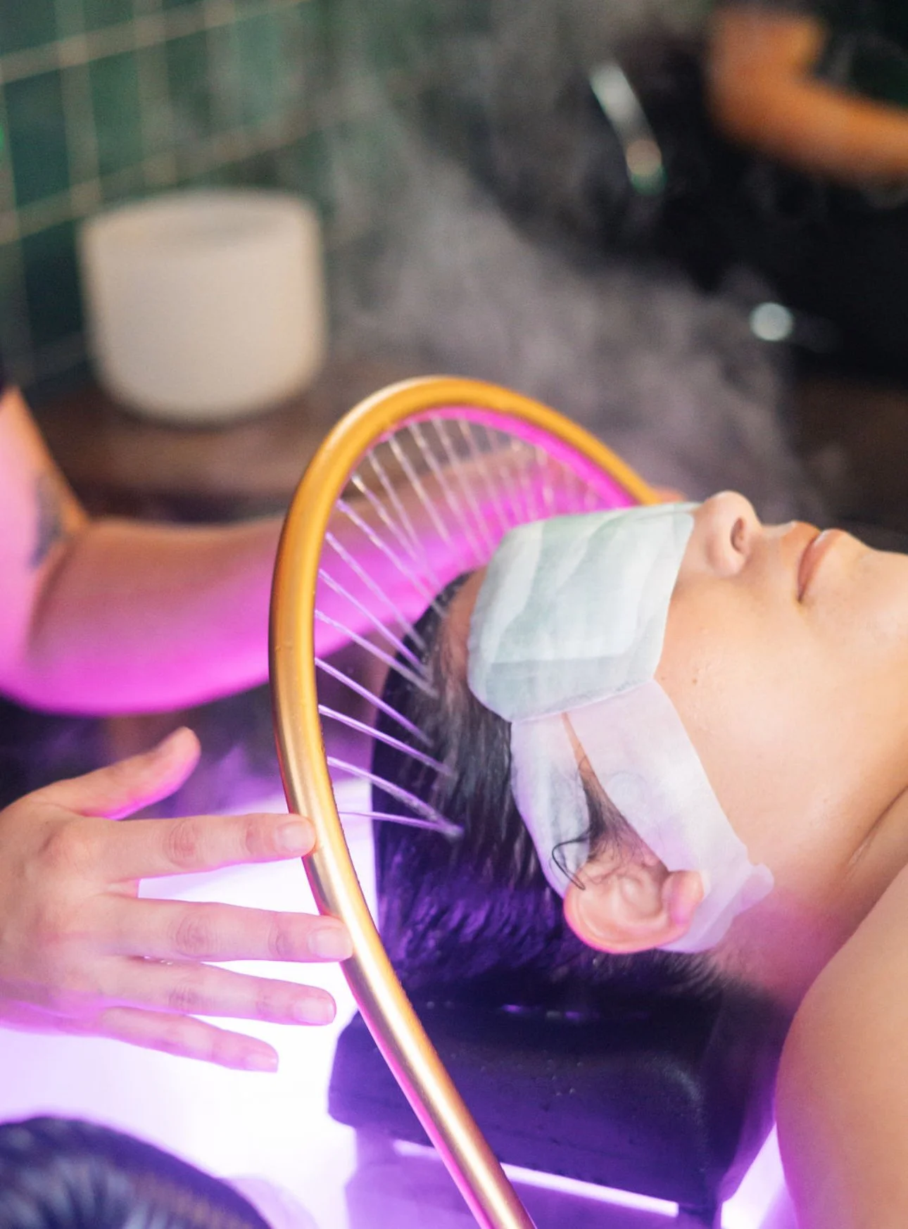 A person receiving a facial treatment with a light therapy device emitting purple light, lying on a treatment bed with a white face mask covering their face, with their eyes closed. Affordable Head spa education for stylists