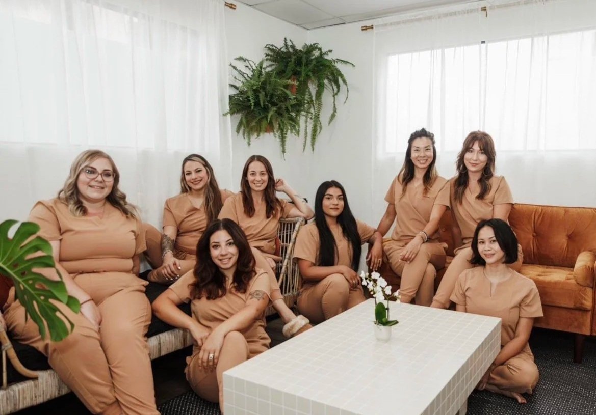 Group of nine women sitting and kneeling in a living room, wearing matching beige outfits, with a coffee table and potted plants in the background. Head spa training.