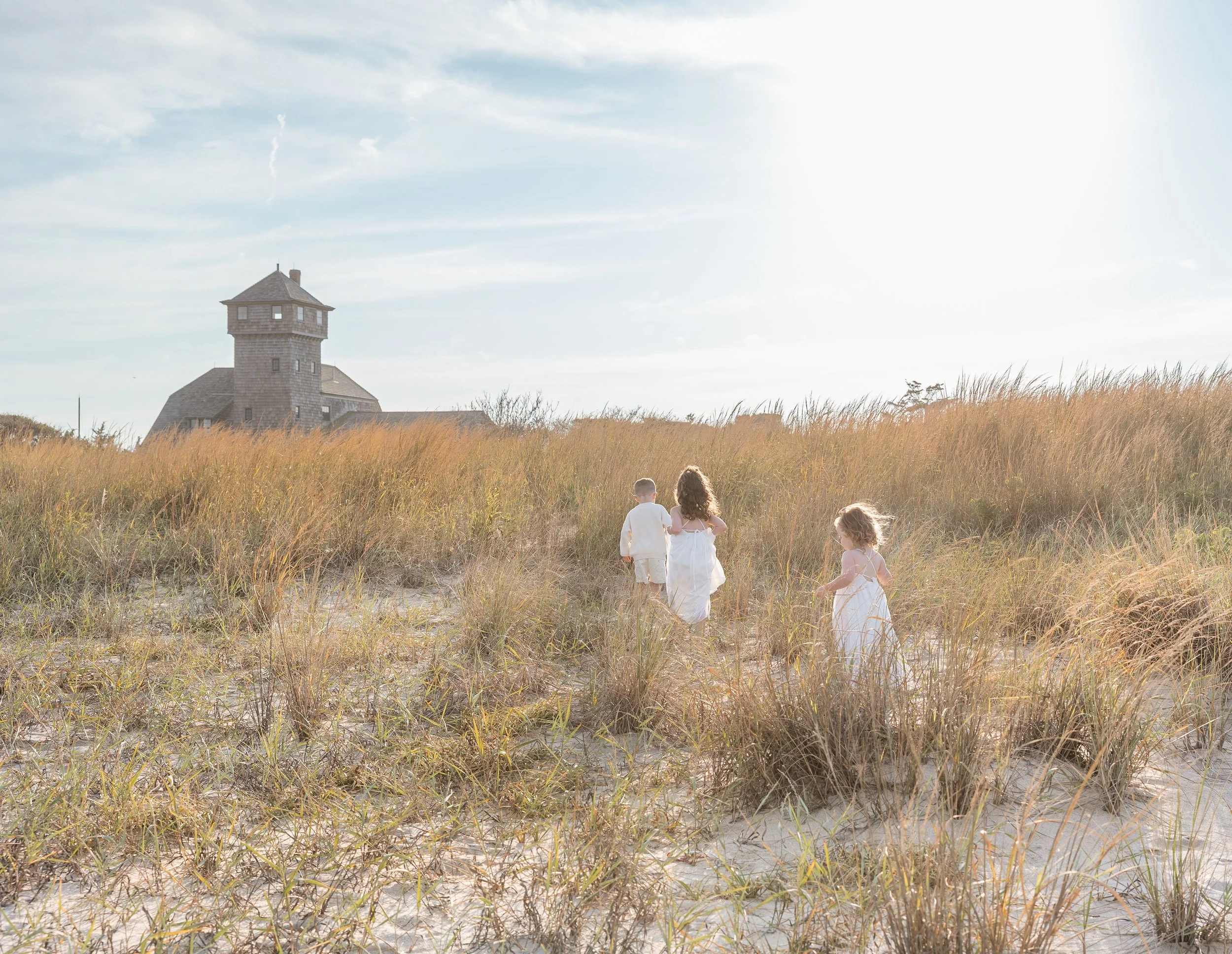 Children walking in a field of tall grass with an old stone building and a tower in the background under a partly cloudy sky.