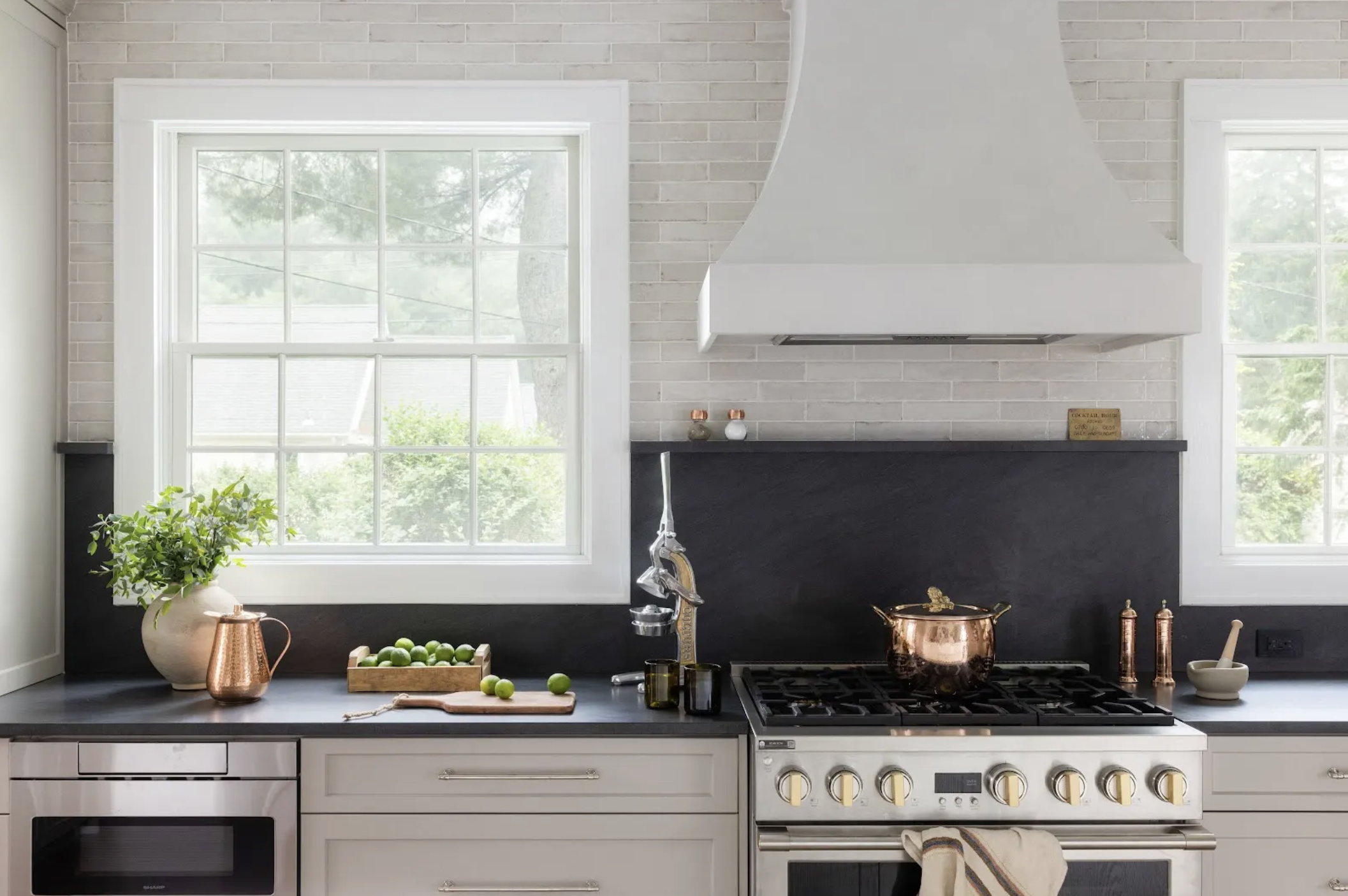A modern kitchen with white cabinets, black countertops, a white brick backsplash, and two large windows. There is a potted plant and copper pitcher on the left, a wooden tray with limes, and various copper and black kitchen utensils on the right.