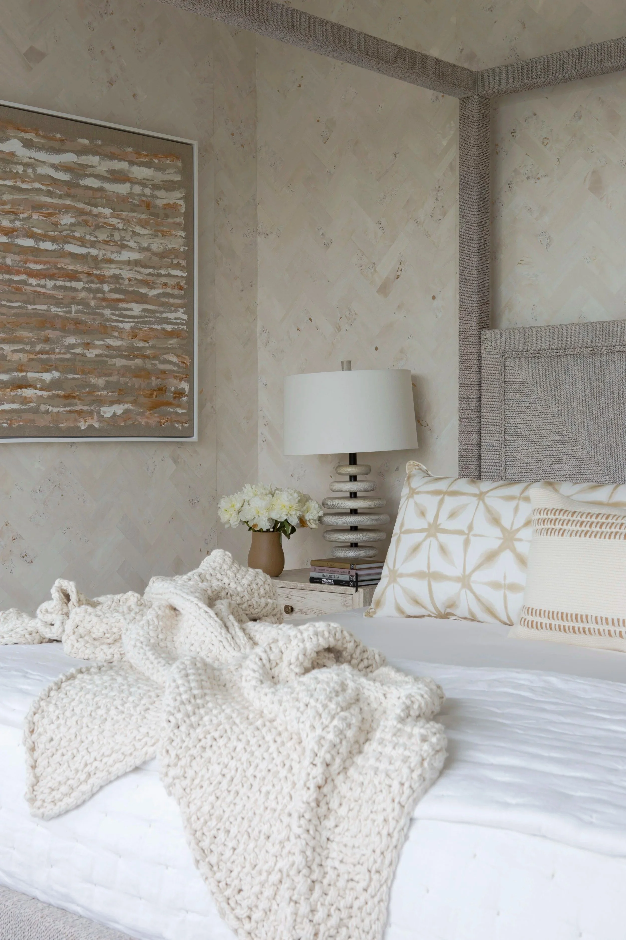 A bedroom with a white bed, a chunky knit beige blanket, decorative pillows in patterned beige and white, a bedside table with a vase of white flowers, a white lamp, and a large abstract artwork on the wall.