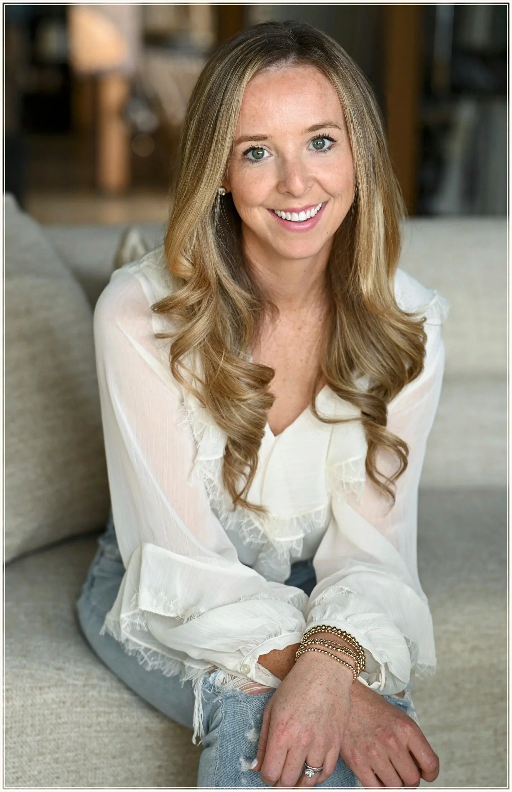A woman with long, wavy blonde hair and green eyes smiling at the camera, sitting on a beige sofa in a well-lit room, wearing a white blouse with lace details, blue jeans, and multiple gold bracelets on her wrist.