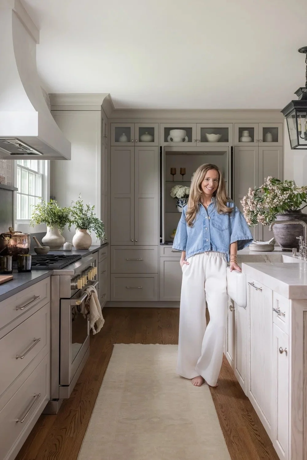 A woman standing in a bright, modern kitchen with grey cabinets, a white countertop, potted plants, and decorative items, smiling and posing casually.