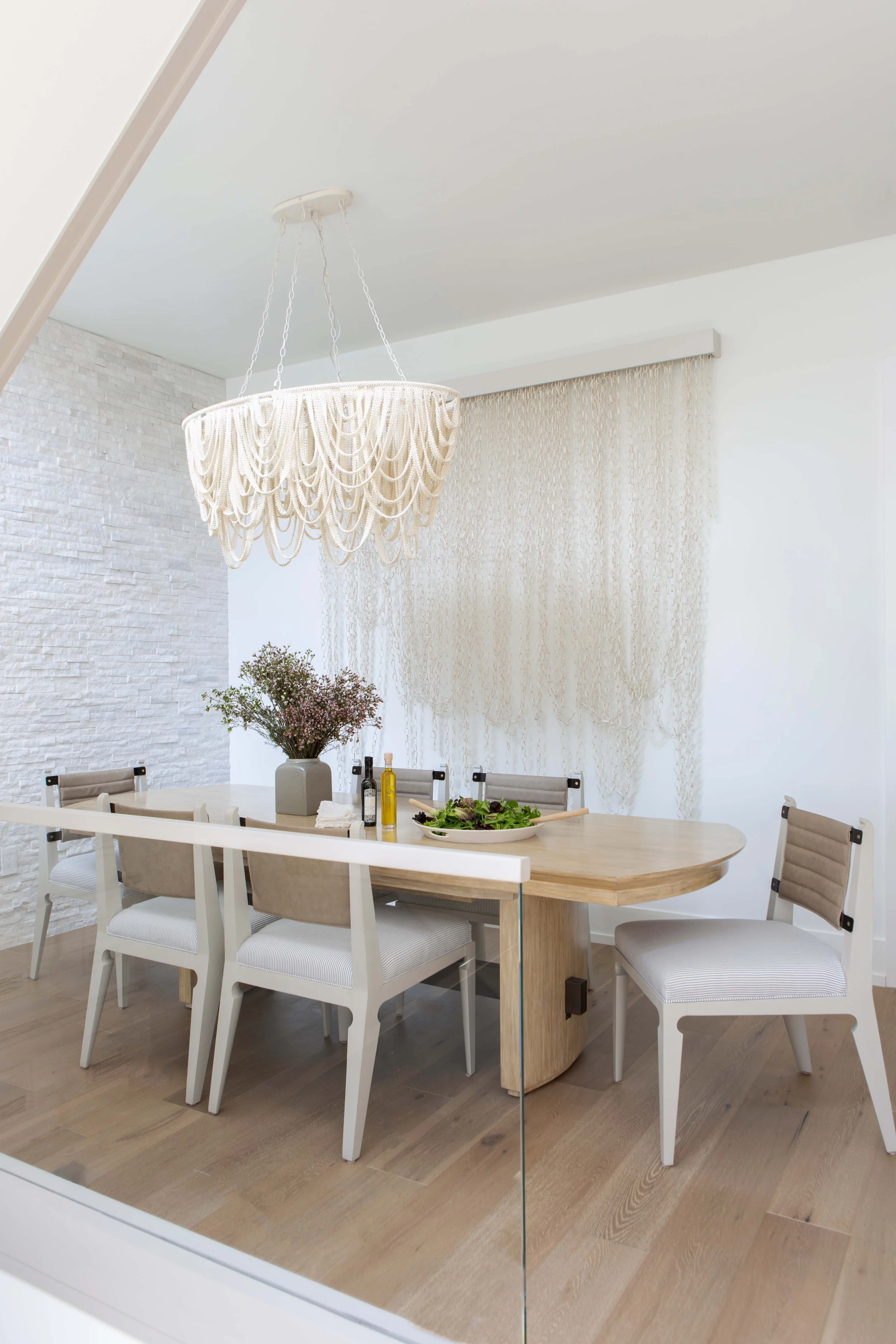 Modern dining room with a light wood table, white chairs, a large hanging chandelier, a white vase with pink flowers, bottles of oil, and a salad, with a textured white brick wall and a window with beige curtains.