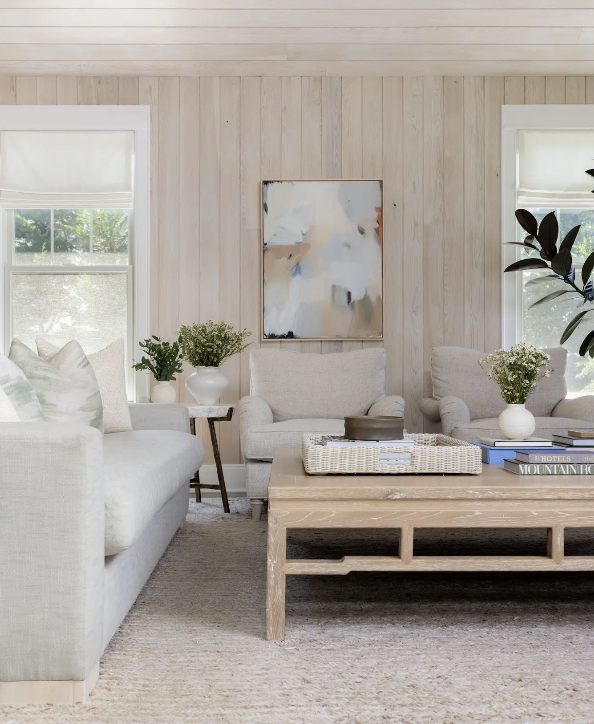 A cozy living room with beige sofas and armchairs, a wooden coffee table, and potted plants. White walls and large windows with blinds allow natural light to fill the space.