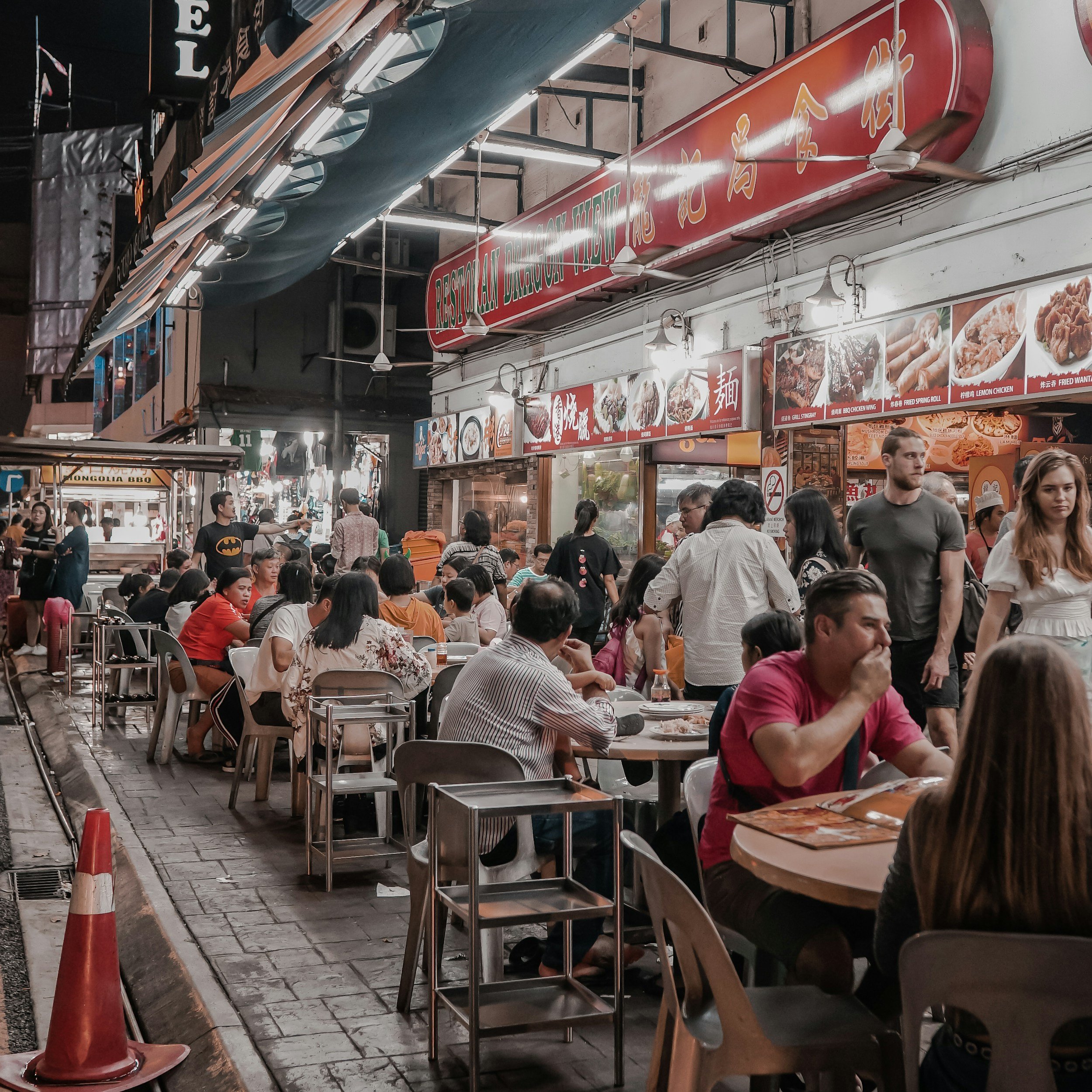 A crowded street food market at night with numerous people sitting at tables and standing around. There are illuminated food stalls with signage displaying menu items and images of various dishes. Overhead lights and a large fan are visible, along with a red sign with Chinese characters.
