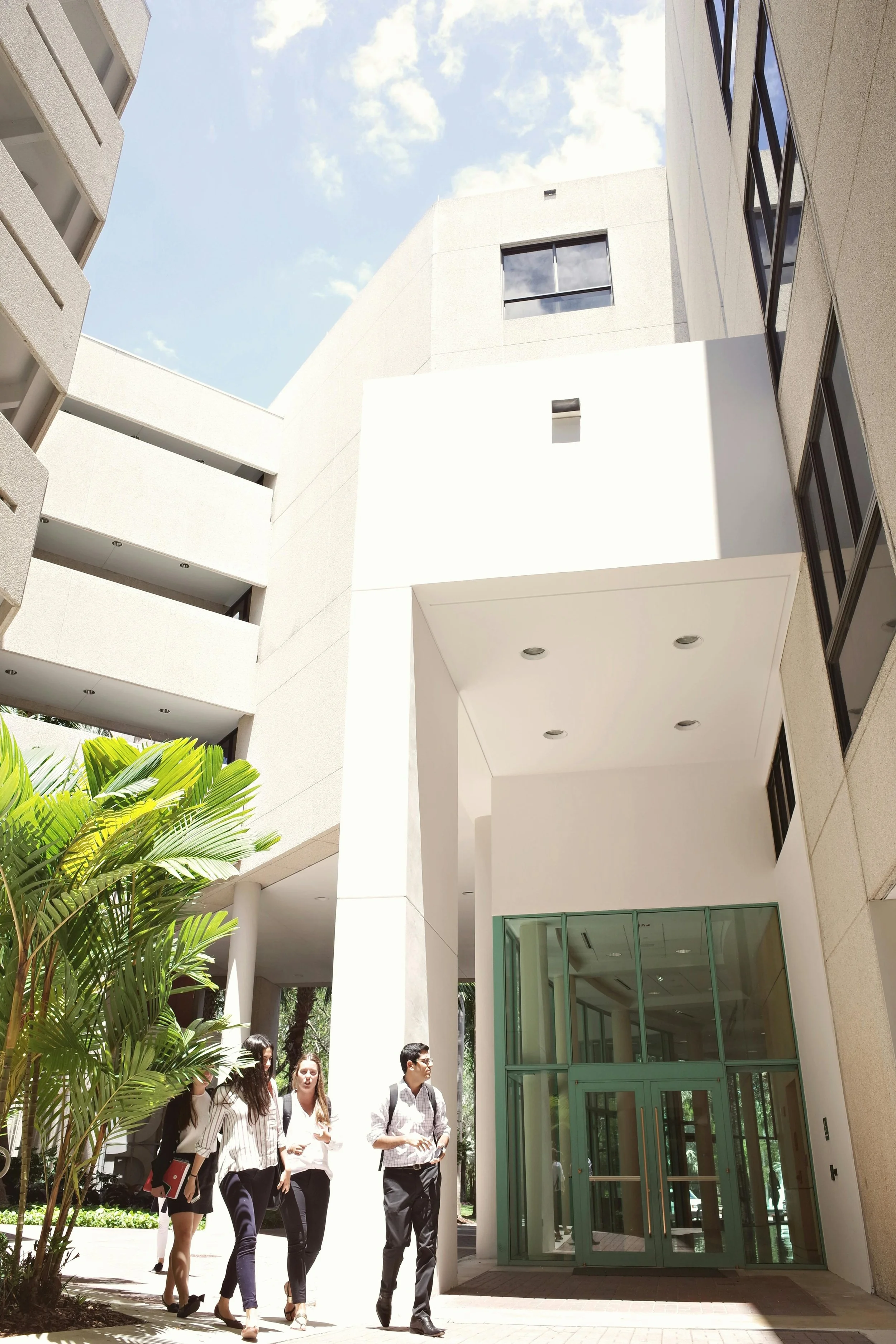 Group of young professionals walking outside a modern office building with white walls, glass doors, and surrounding greenery on a sunny day.