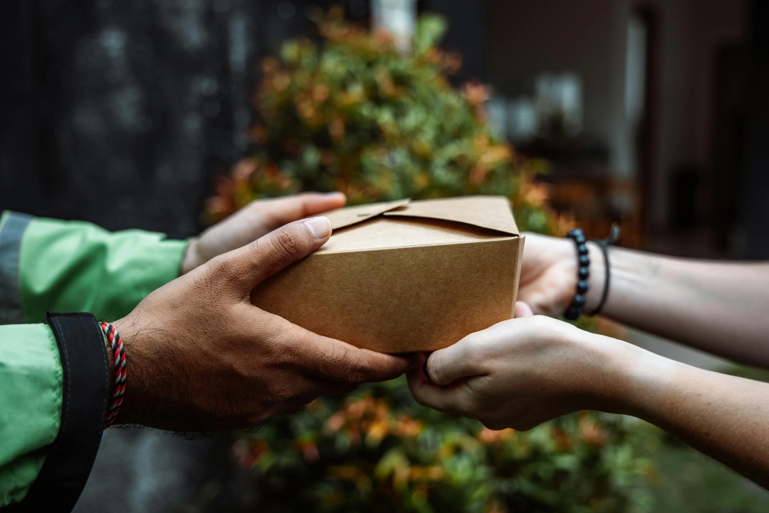 Multiple hands exchanging a brown paper takeout box outdoors, with blurred colorful autumn leaves and a building in the background.