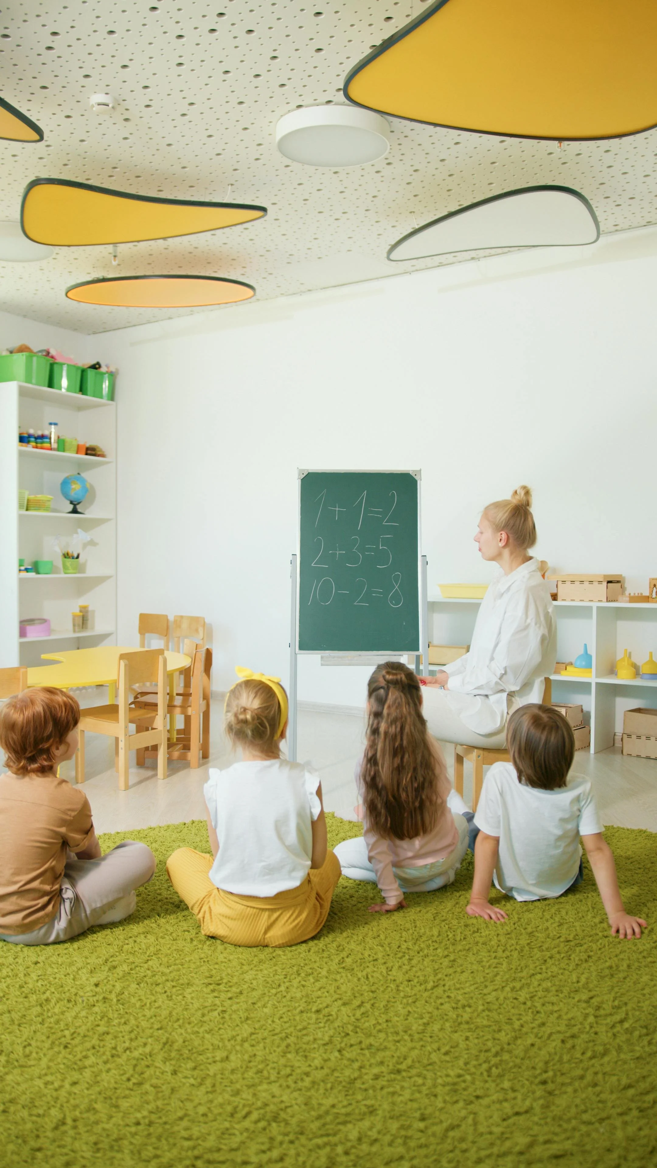 Children sitting on a green rug facing a teacher in a classroom with a blackboard displaying simple math problems.