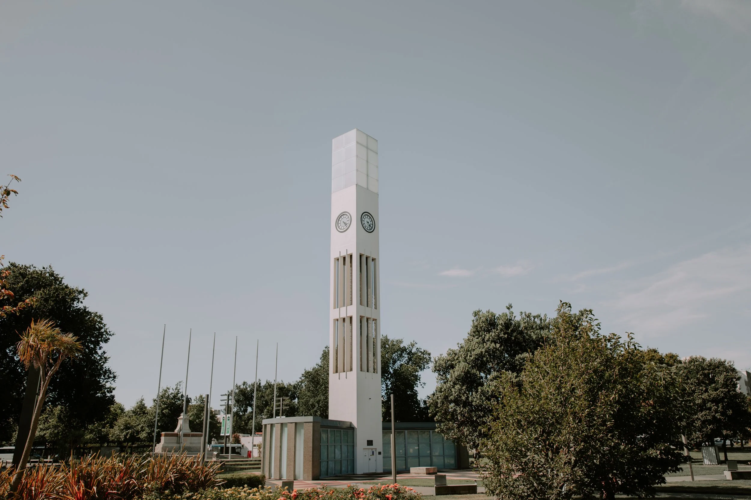 A tall white clock tower with two clocks, set in a park surrounded by trees and a clear sky.