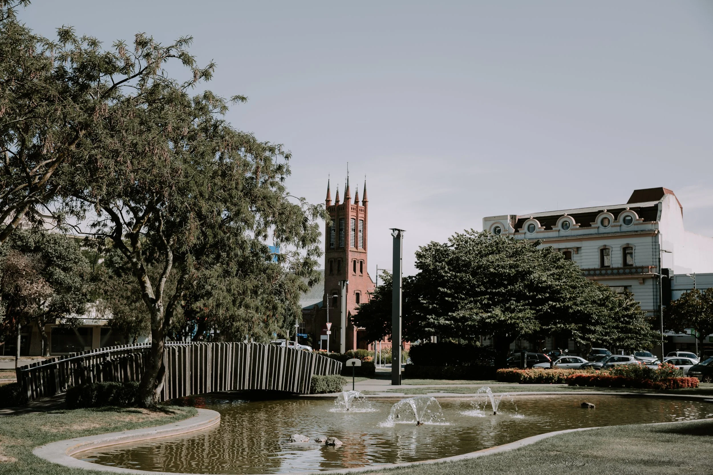 A park with a pond and fountains, surrounded by trees and benches, with historic buildings and a church in the background under a clear sky.
