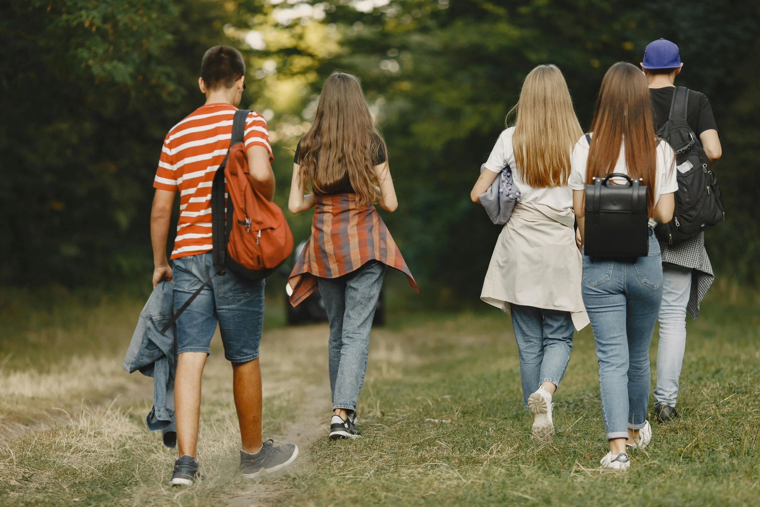A group of five young people walking on a grassy trail in a forest, seen from behind, all carrying backpacks.