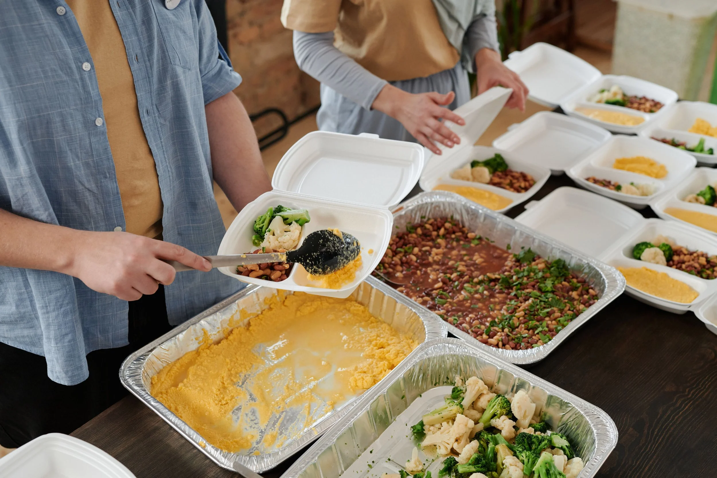 People serving food at a buffet with various dishes including cauliflower, broccoli, beans, and yellow rice.