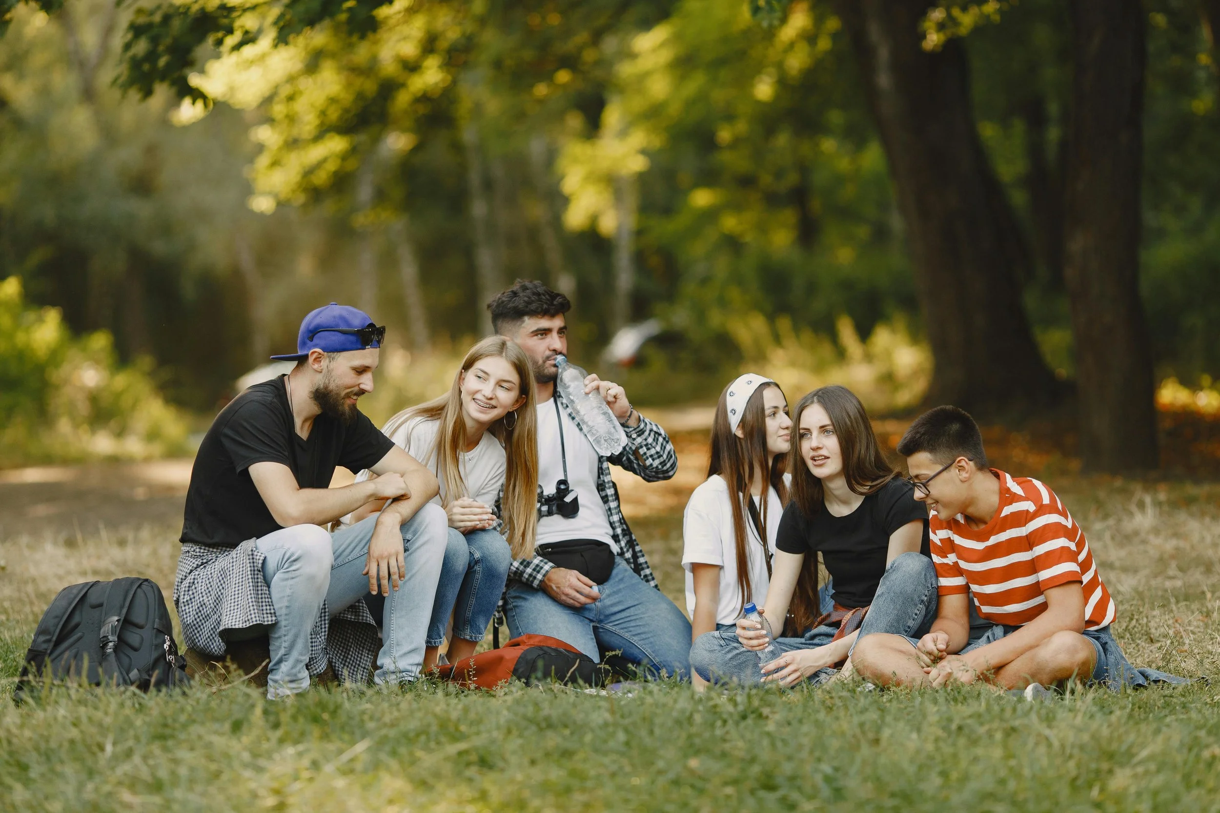 A group of young friends sitting on grass in a park, engaging in conversation and smiling, with trees and a river in the background during autumn.