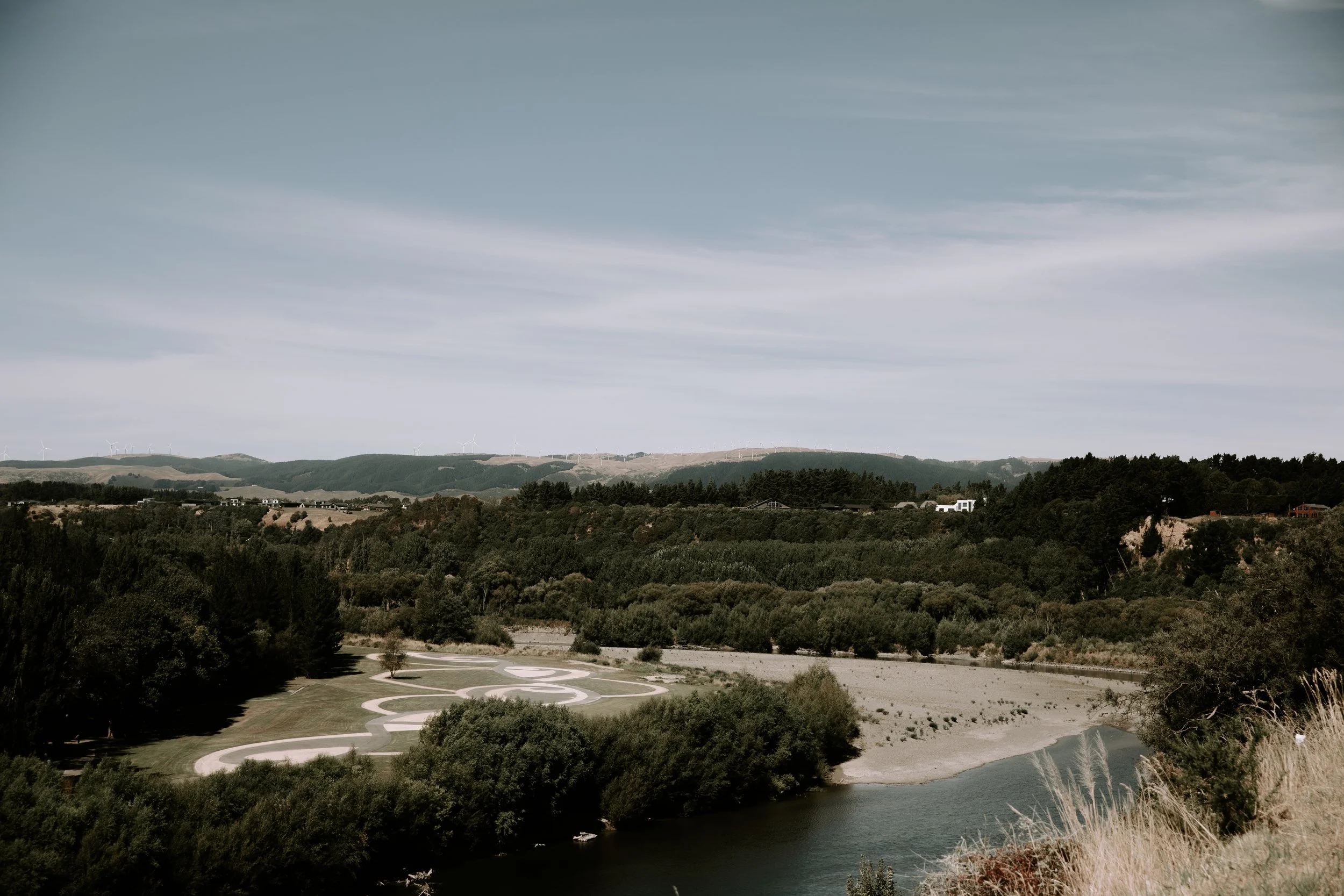 A landscape view of a river winding through a lush green area with a lawn and a distinctive winding paved path, set against rolling hills with wind turbines and a cloudy sky overhead.