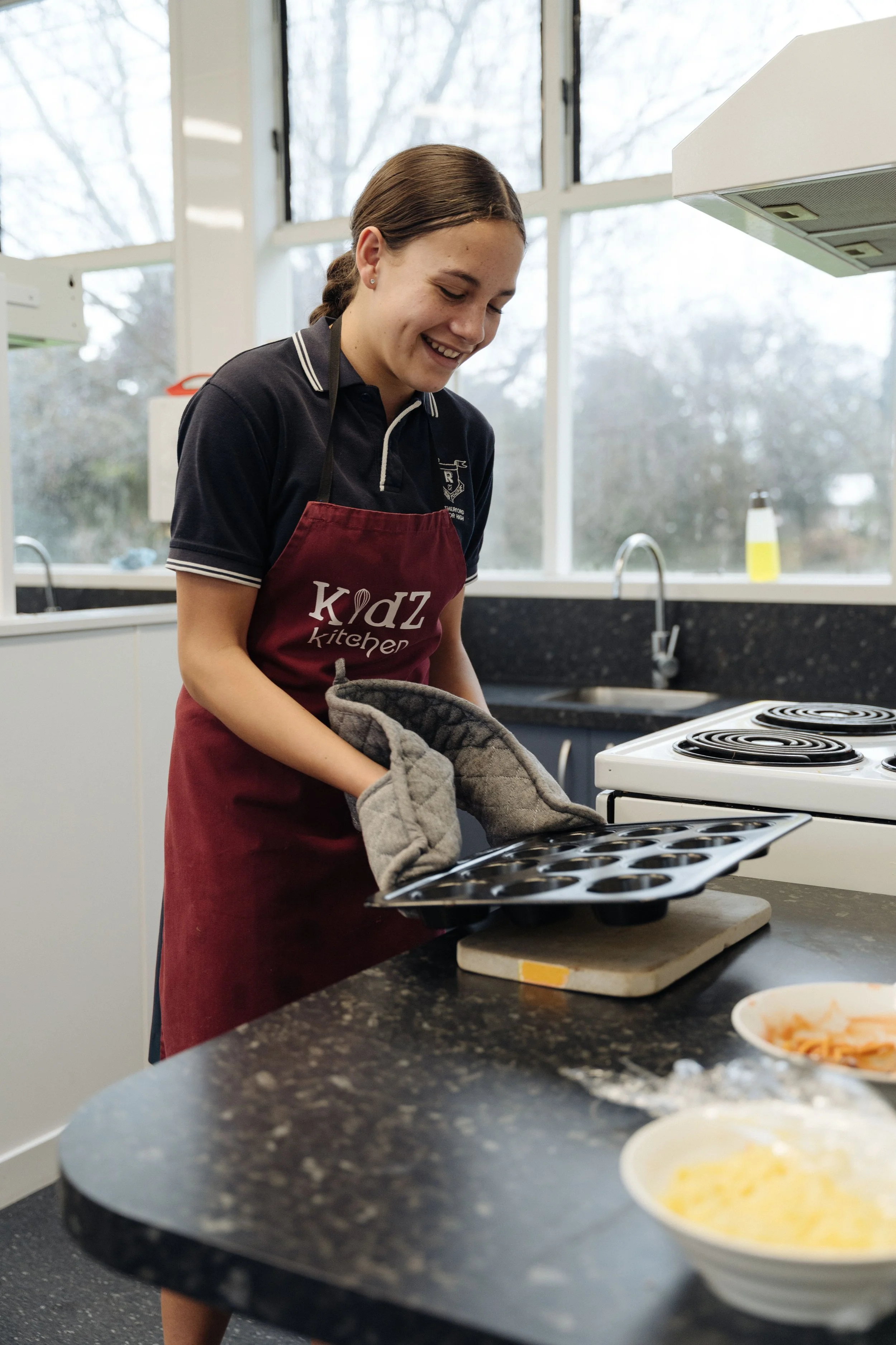 A young girl with brown hair in a braid, wearing a maroon apron with 'KIDZ Kitchen' written on it, is smiling and holding a baking tray in a kitchen. There are bowls of food on the counter and a window in the background.