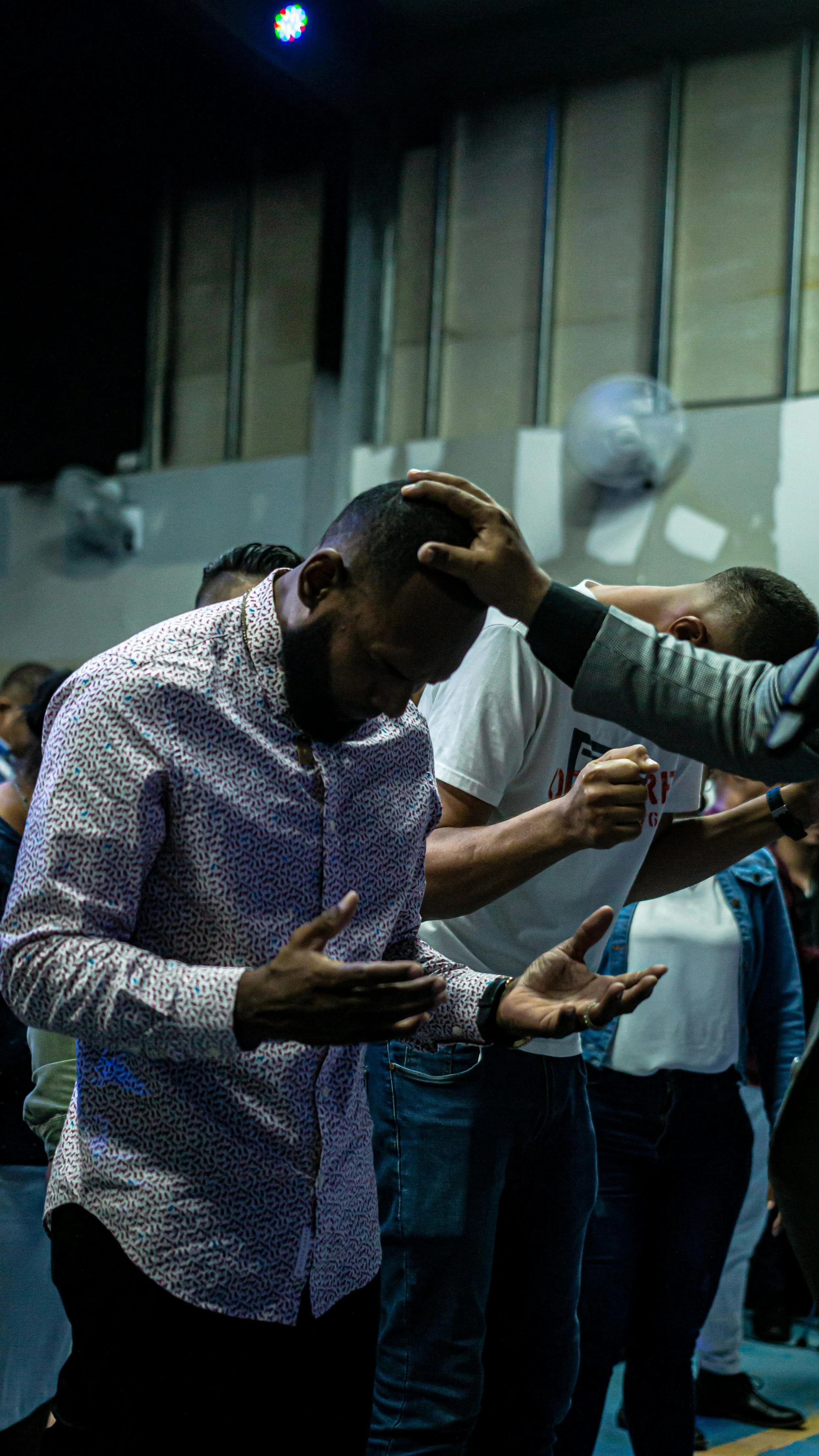 People standing with hands raised and bowed heads, engaged in prayer or worship in an indoor setting.