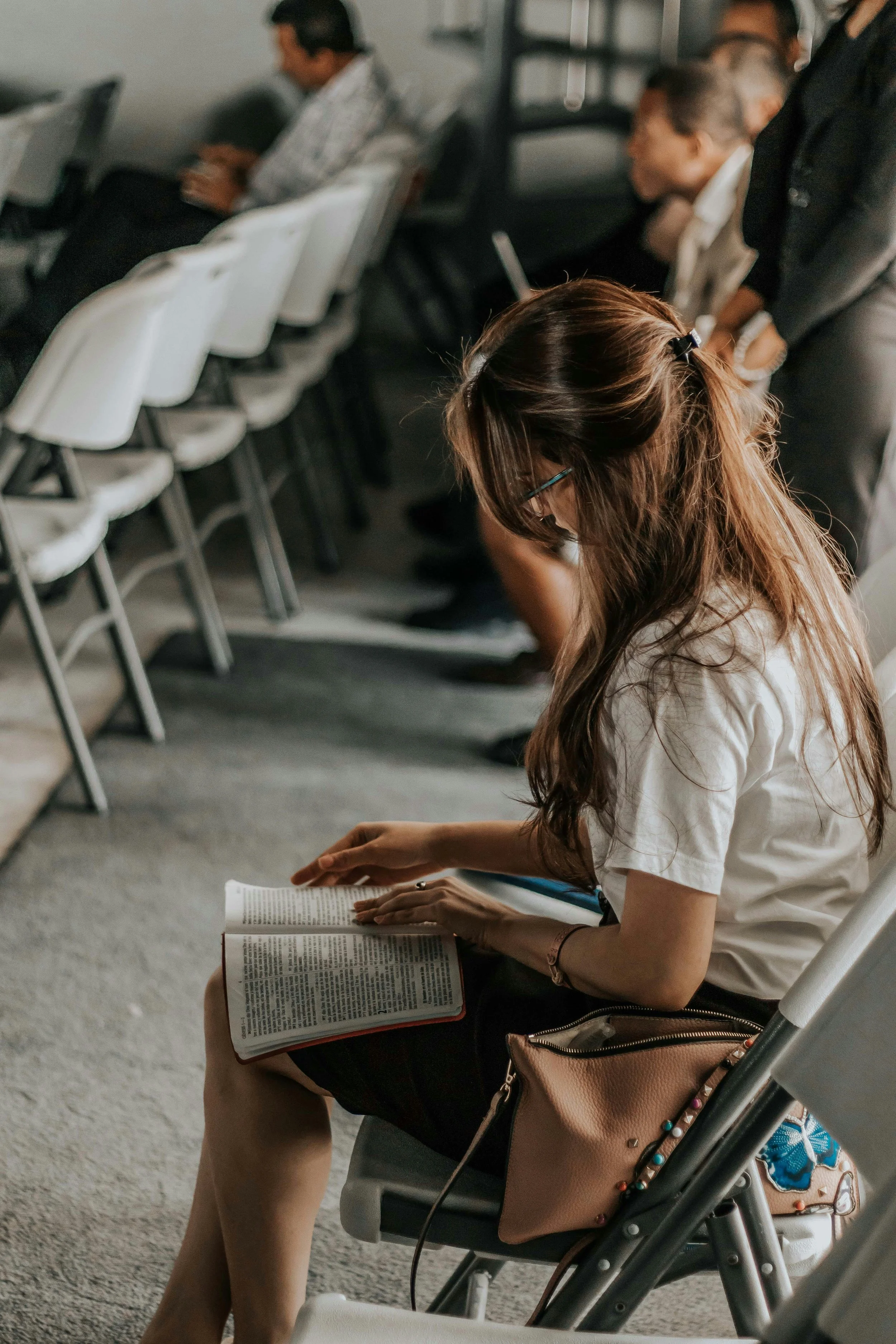 A woman with long brown hair and glasses sitting in a row of chairs in what appears to be a waiting area, reading a book or magazine. She has a tan purse with colorful embellishments resting on her lap and is wearing a white shirt.