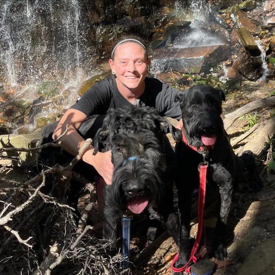 Ann, Pet Groomer at Tails and Ales in Hickory, NC, crouches by a waterfall with her two Giant Schnauzers as they take a break during a hike for a picture.