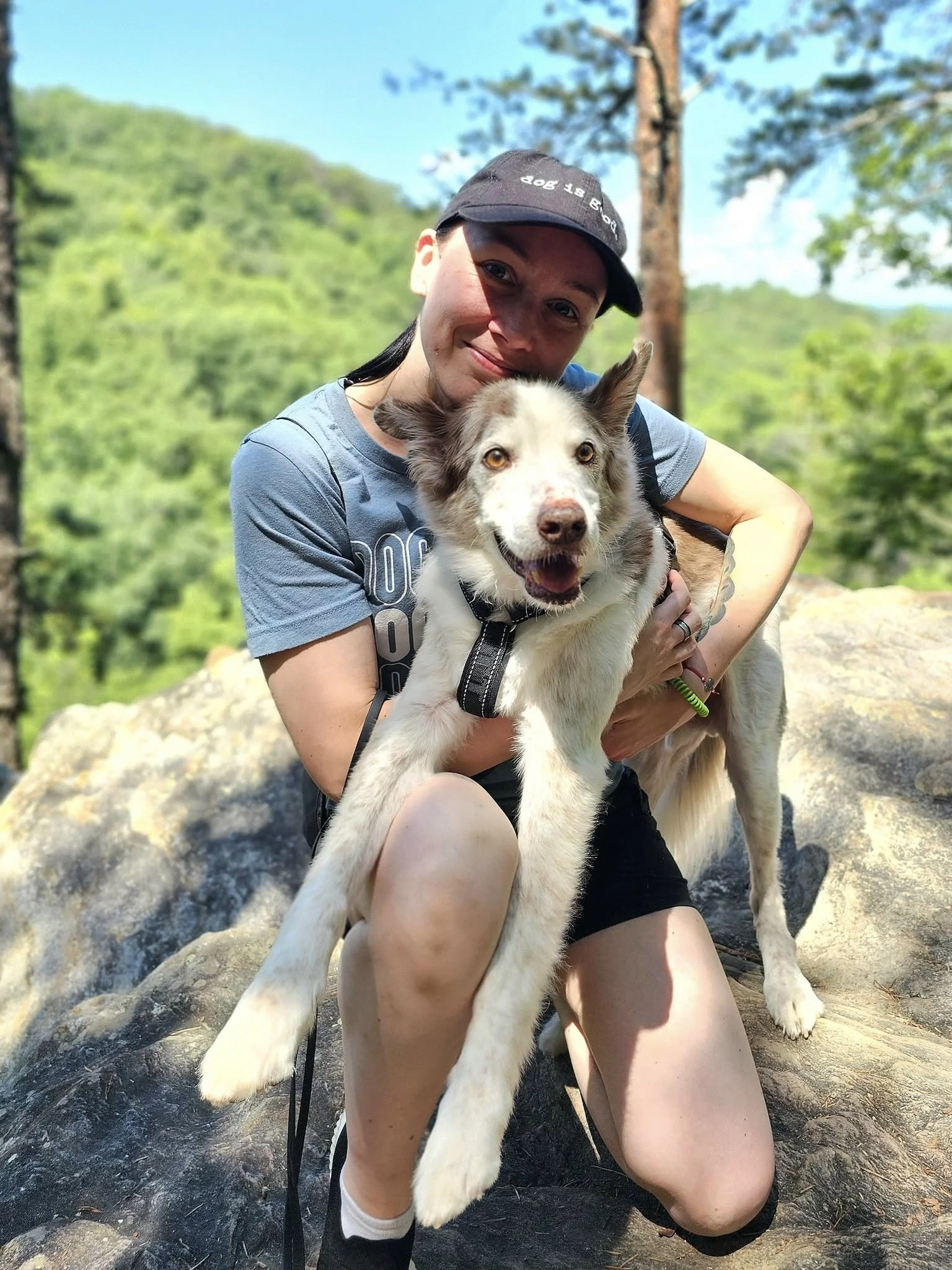 Maggie, Pet Groomer at Tails and Ales in Hickory, NC, crouches on a rock and holds her dog as they take a break during a hike for a picture.