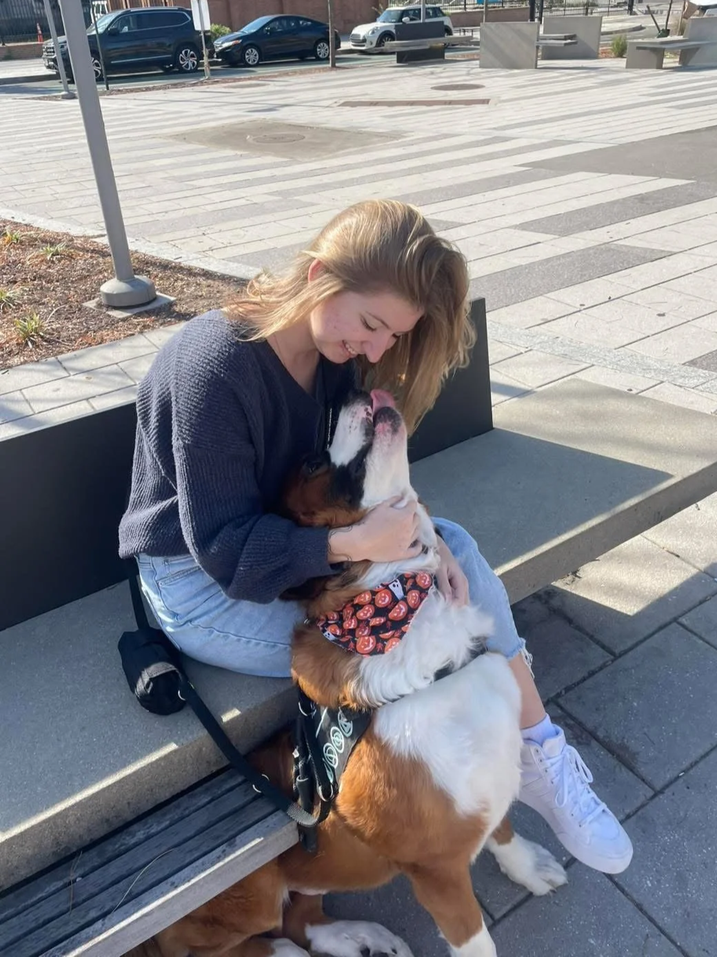 Skylar, Pet Groomer at Tails and Ales in Hickory, NC, sits on a bench and smiles down toward her large mixed breed brown and white dog who looks back up at her and tries to lick her face.