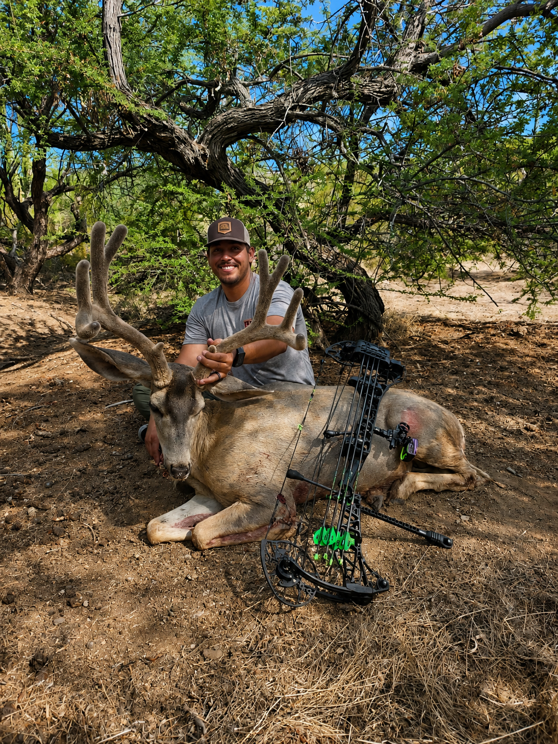 A man smiling and kneeling next to a freshly hunted deer with large antlers, in a wooded outdoor area. The deer has a bow and arrows attached to it.