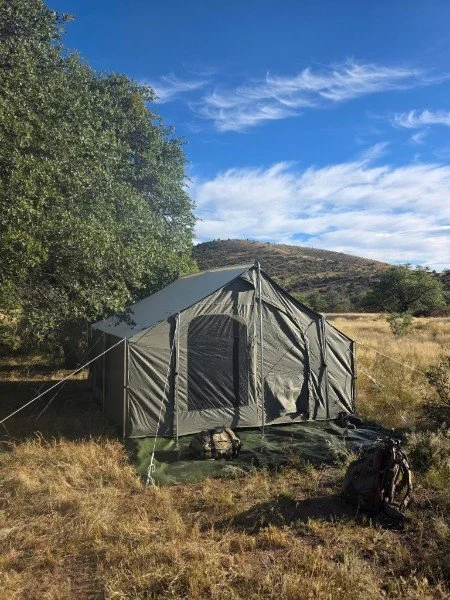A camping tent set up in a grassy field with a backpack nearby, surrounded by trees and hills under a blue sky with some clouds.