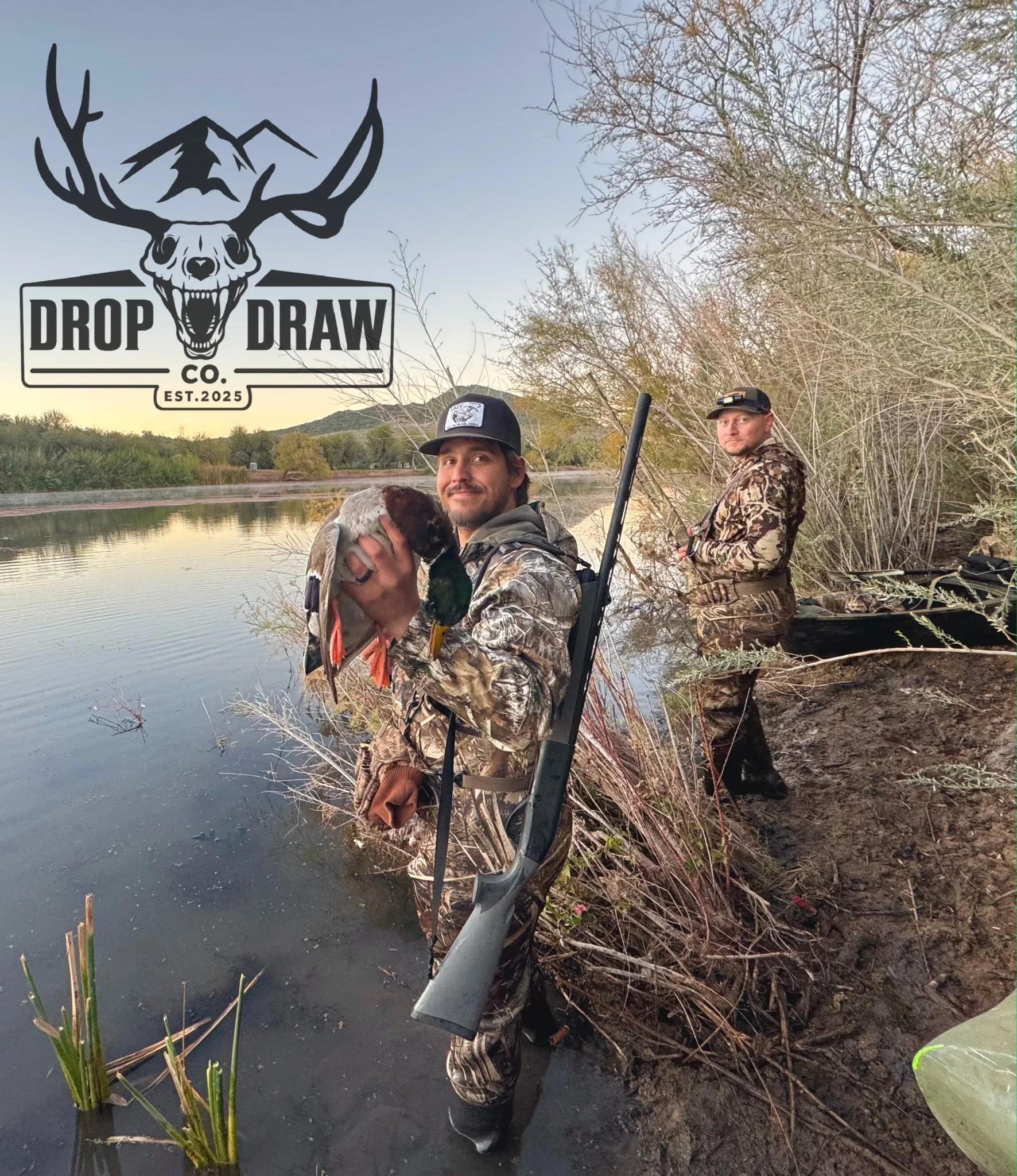Two hunters in camouflage clothing are standing by a riverbank with a boat and a shotgun. One hunter is holding a dead duck, and the other is standing nearby. There is a logo with a deer skull and the text "Drop Draw Co. Est. 2025" in the top left co
