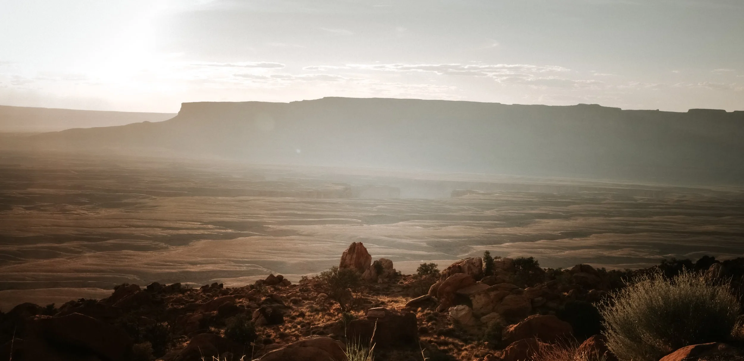 A vast desert landscape with layered rock formations and mesas in the distance, under a cloudy sky during sunset or sunrise.