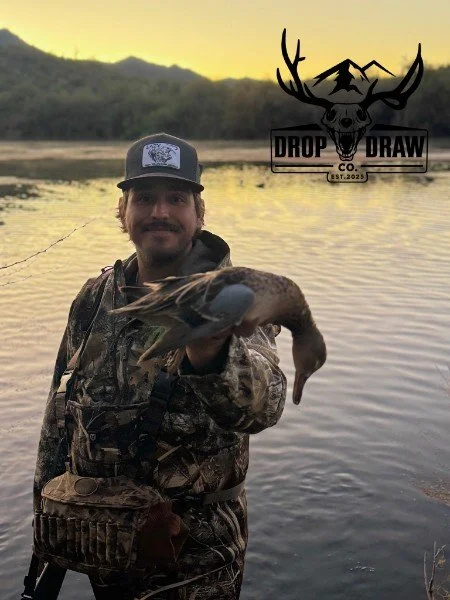 A man standing by a lake at sunset, holding a large fish he caught, wearing camouflage clothing and a baseball cap, with a logo for Drop Draw Co. featuring a deer skull with antlers in the top right corner.