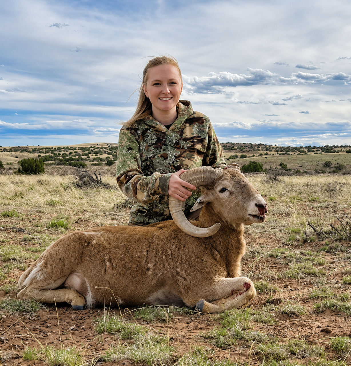 A young woman in camouflage clothing kneels on grassy terrain, smiling, while holding the horns of a freshly hunted ram with light brown fur and curved horns, lying on the ground in an open landscape under a partly cloudy sky.