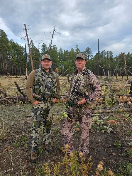 Two men in camouflage clothing stand outdoors in a forested area; one holds a bow, and both are smiling.