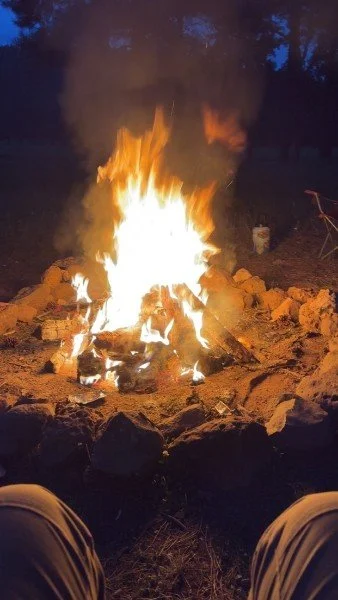 Campfire burning with flames and smoke in a rocky outdoor area at night.