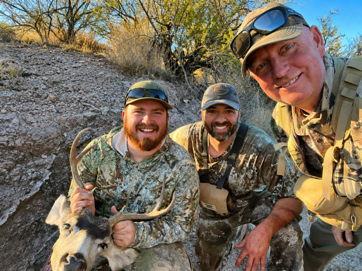 Three men in camouflage clothing smiling and posing for a photo with a deer they have hunted, outdoors with dry trees and bushes in the background during daylight.