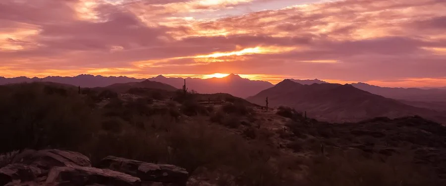Sunset over a desert mountain landscape with cactus and rocky terrain.