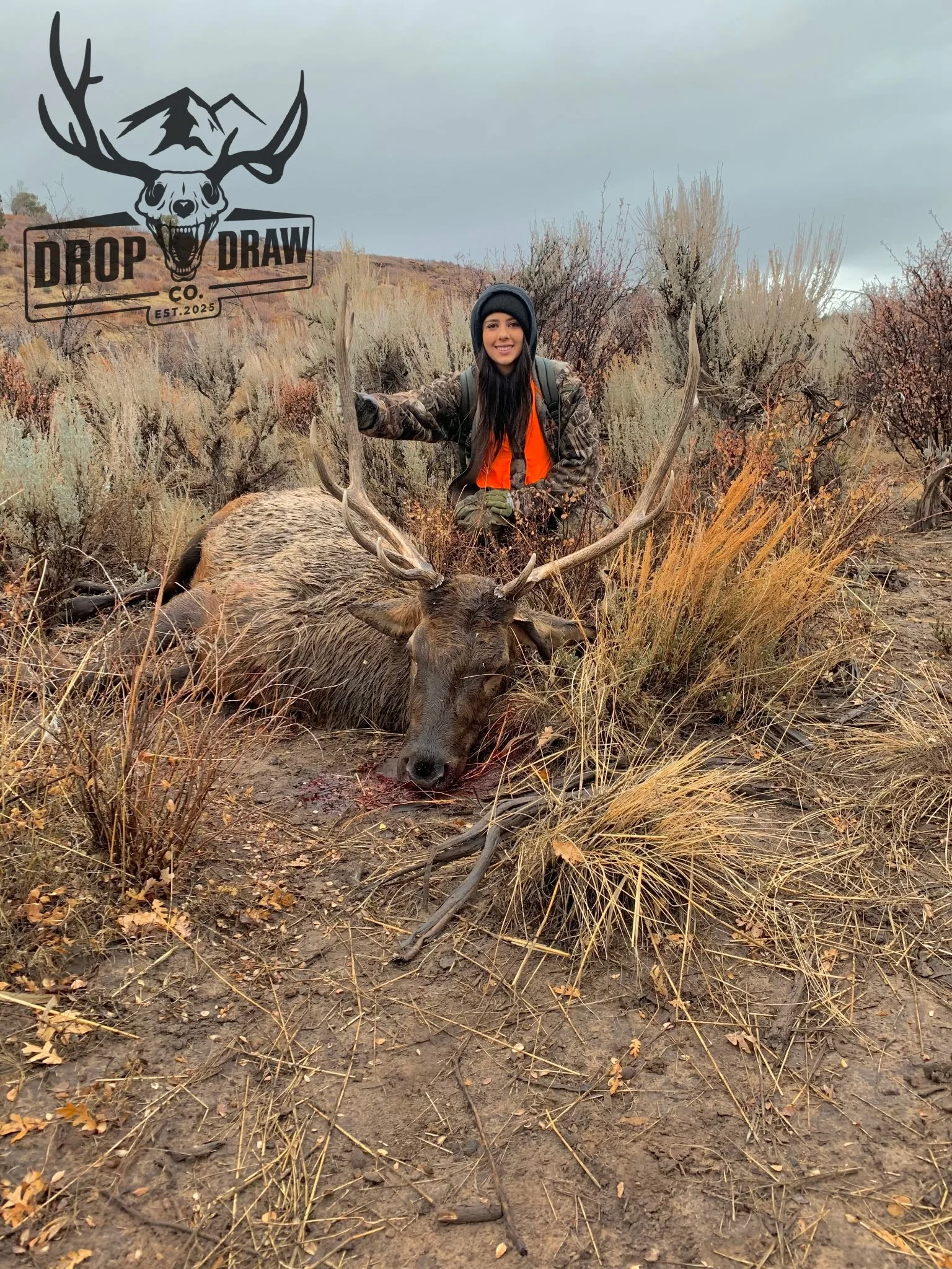 A young woman in camouflage clothing and orange vest posing with a large deer carcass in a dry, shrubland area with overcast sky.