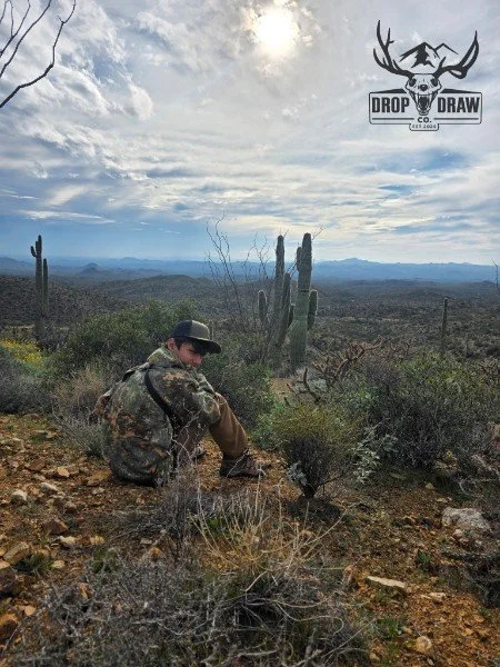 A person sitting on rocky desert terrain surrounded by cacti and desert shrubs, with a cloudy sky overhead and mountains in the distance. There is a logo of a deer skull with antlers and the text 'Drop Draw Co' in the top right corner.