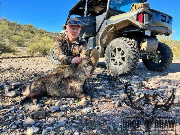 A man in outdoor gear kneels next to a large dead wild boar on rocky ground, with a black off-road vehicle in the background, under a clear blue sky.