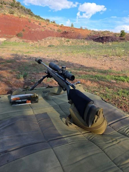 A rifle with a scope is resting on a shooting mat outdoors, with a box of target pellets nearby, on a field with colorful hills and a blue sky in the background.