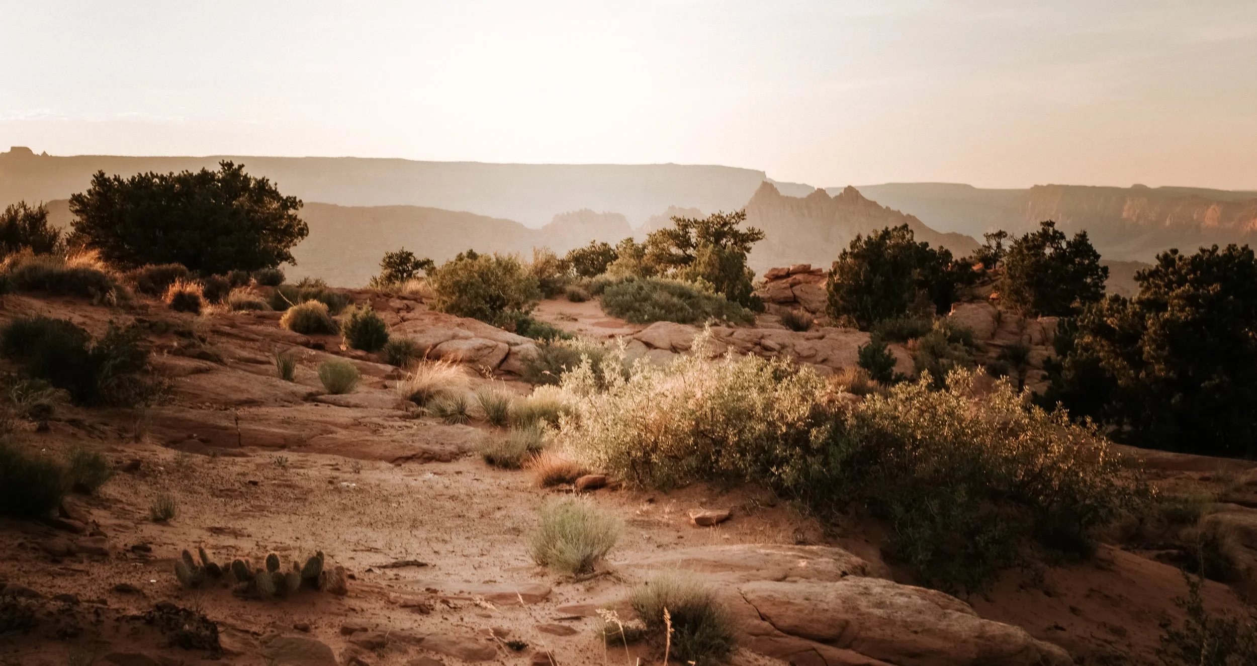 Sunset over a desert landscape with rocky terrain, sparse bushes, and distant cliffs.