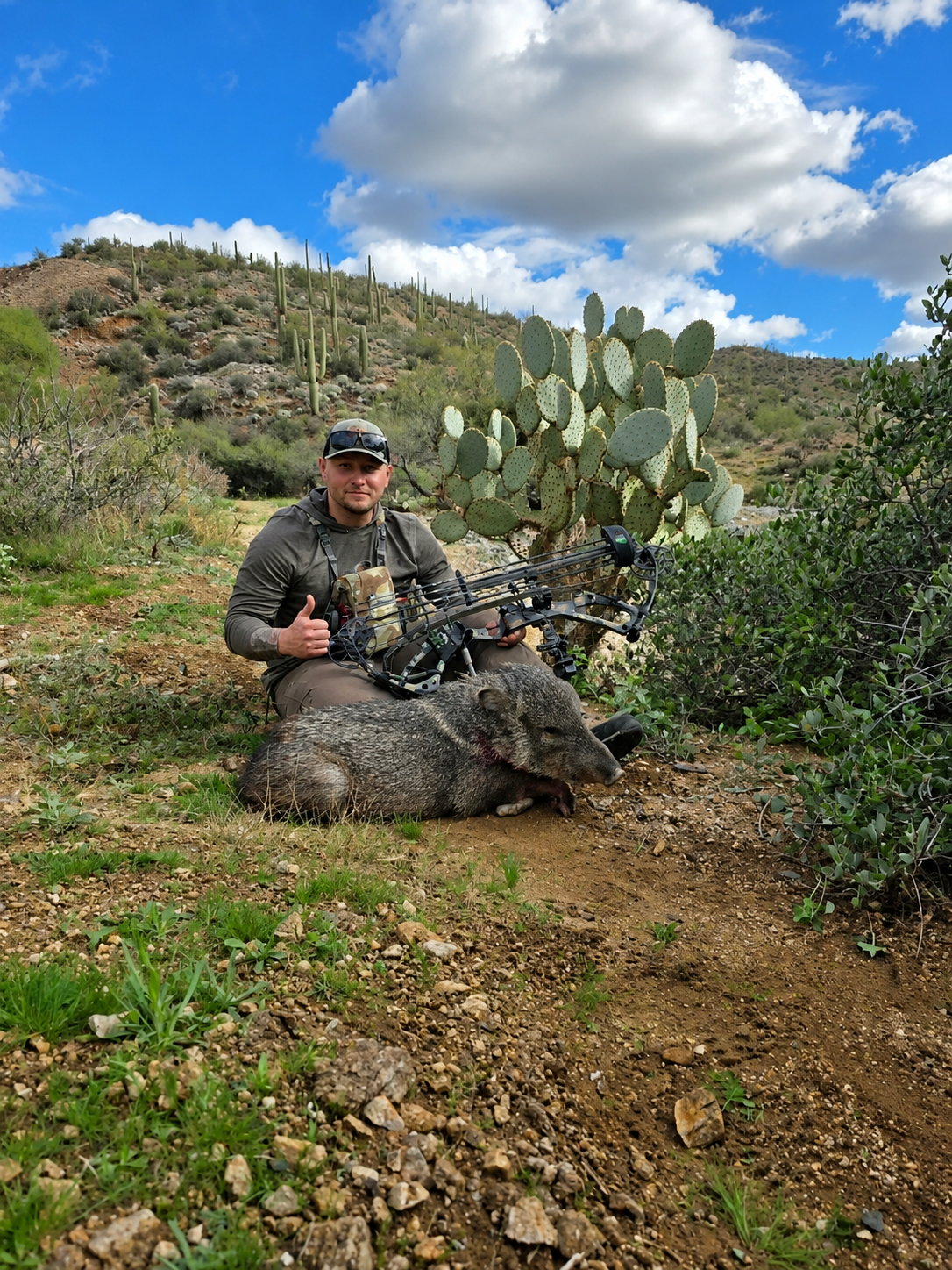 A man outdoors in a desert landscape with cacti, holding a bow, and posing next to a dead wild boar.