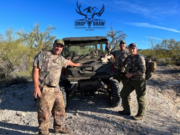 Three men in camouflage clothing standing outdoors next to a black off-road vehicle with a mounted deer skull, in a desert-like landscape with sparse bushes and trees, under a clear blue sky, with a Drop and Draw logo visible in the sky.