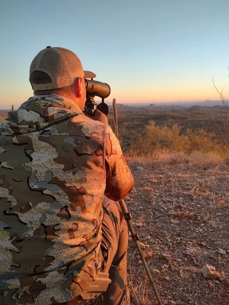 A person in camouflage clothing and a cap using a rifle with a scope, sitting in a desert landscape during sunset.